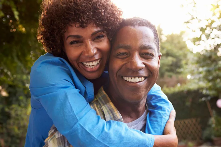 Autoimmune-Doctor A middle-aged African American couple outside, the woman embracing the man from behind pressing her cheek to his, both are smiling due to successful Autoimmune Disorder treatment from Frank Lanzisera, D.C. in Tampa Bay Metro.