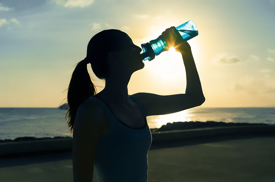 Chelation-and-Detoxification-Clinic A woman in exercise clothes drinking from a water bottle on the beach, with the water and sunset in the background. Get chelation and detoxification treatment from Frank Lanzisera, D.C. in Tampa Bay Metro.
