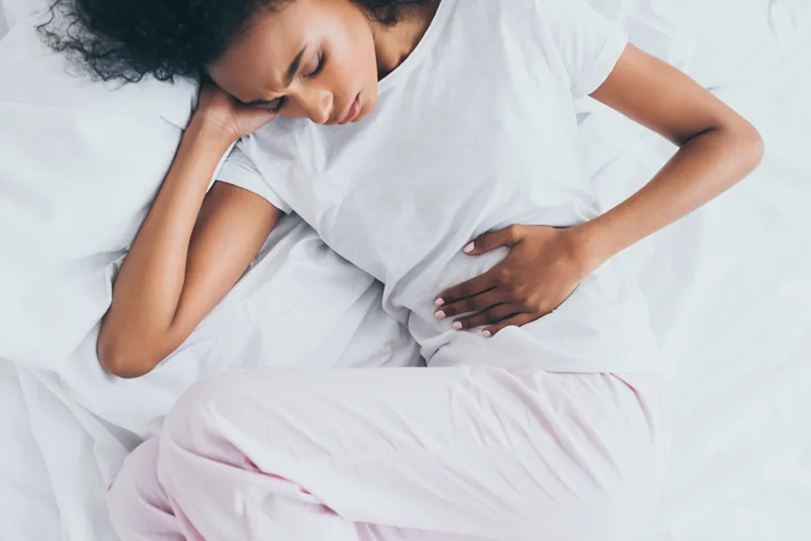Crohns-Disease-Clinic A dark-skinned woman in white clothes lays on her bed clutching her middle before getting treatment for Crohn's Disease from Frank Lanzisera, D.C. in Tampa Bay Metro.