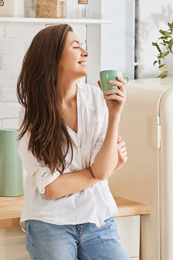 Crohns-Disease-Treatment A woman with a white blouse an jeans drinking tea in her kitchen, smiling after getting treatment for Crohn's Disease from Frank Lanzisera, D.C. in Tampa Bay Metro.