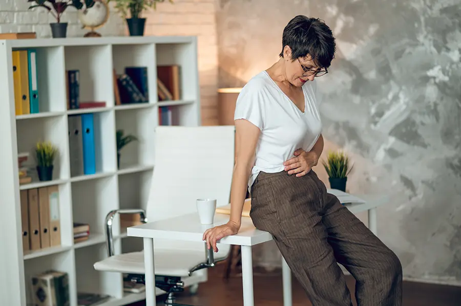 Digestive-IV-Therapy-Clinic A middle-aged woman leans against her desk holing her belly in pain. Schedule IV Therapy for digestive health from Frank Lanzisera, D.C. in Tampa Bay Metro.