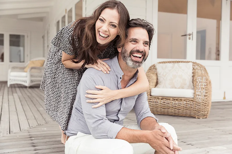 Health-and-Wellness-Services-Doctor A middle-aged couple on a wooden porch with wicker furniture; the woman is playfully hugging the man from behind. They are benefiting from the health and wellness services from Frank Lanzisera, D.C. in Tampa Bay Metro.