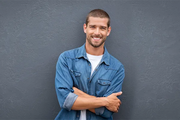 Hormones-for-Men-Doctor A man in a denim shirt stands smiling against a gray-blue wall, pleased with his testosterone hormone therapy from Frank Lanzisera, D.C. in Tampa Bay Metro.