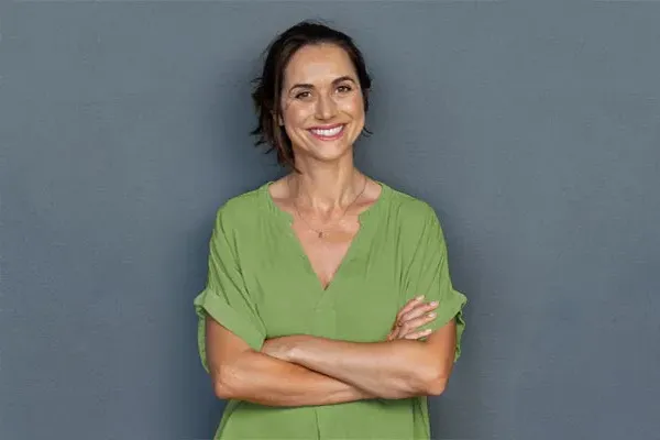 Hormones-for-Women-Doctor A woman in a green shirt stands smiling against a gray-blue wall, pleased with her hormone therapy from Frank Lanzisera, D.C. in Tampa Bay Metro.