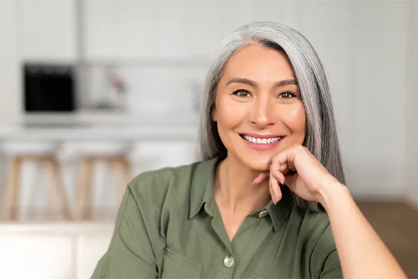 Menopause-Clinic An asian woman with long gray hair and a green shirt sitting and smiling on her couch, happy with her menopause care from [NAME} in Tampa Bay Metro.