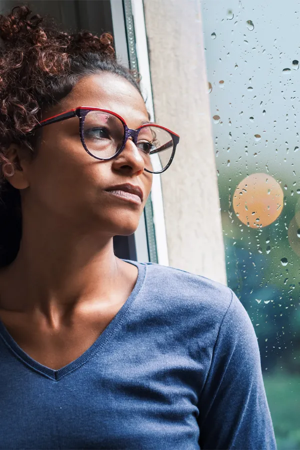 PMDD-Treatment A black woman with with a somber expression and glasses and a gray shirt stares out the window on a rainy day. Get treatment for Premenstrual Dysphoric Disorder from Frank Lanzisera, D.C. in Tampa Bay Metro.