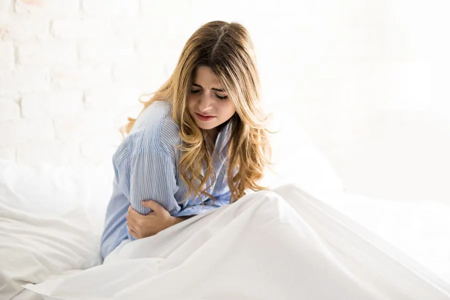 PMS-Therapy A woman with long blonde hair sits in her bed holding her stomach in pain, considering getting care for PMS from Frank Lanzisera, D.C. in Tampa Bay Metro.