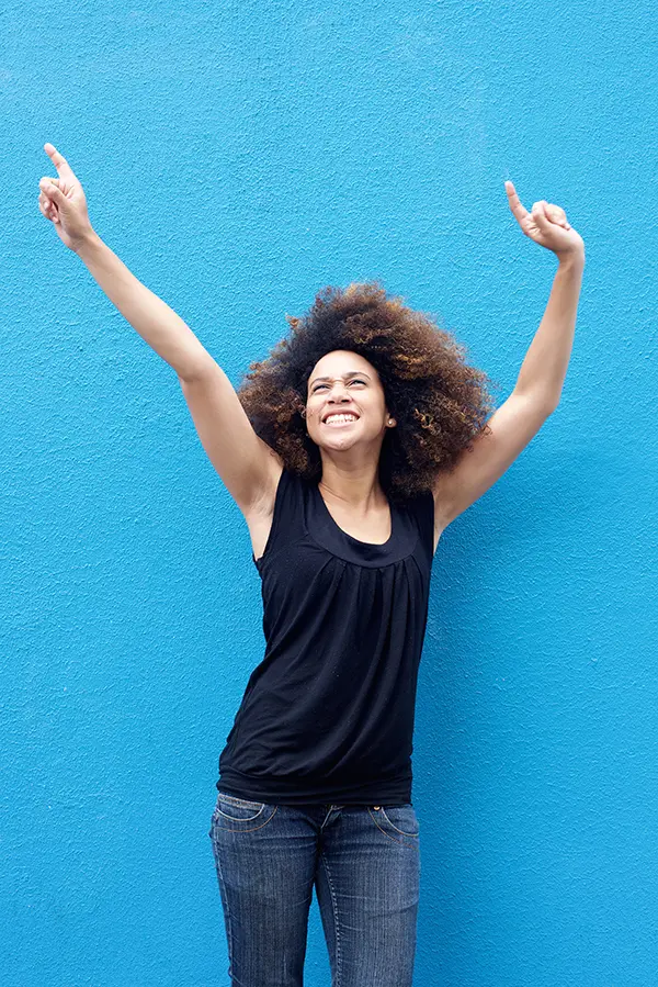 PMS-Treatment A woman in a dark blue tank top standing in front of a bright blue wall, raising her arms in celebration of relief from PMS from Frank Lanzisera, D.C. in Tampa Bay Metro.
