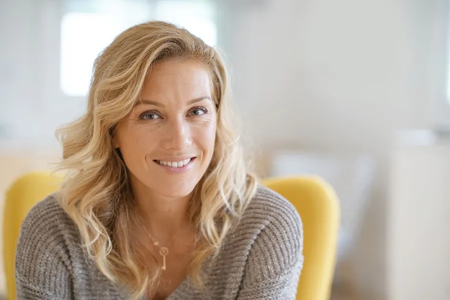 Perimenopause-Doctor A blonde woman wearing a a gray sweater sits inside in a yellow chair, smiling and happy with her perimenopause treatment from Frank Lanzisera, D.C. in Tampa Bay Metro.